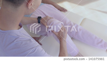 Supporting lilac-shirt caregiver guiding senior's bent knee in pink sweats home on sofa, smartwatch 141075981