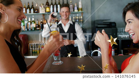 Laughing women at bar wearing dark and red dresses holding cocktails, bartender shaking shaker 141076145
