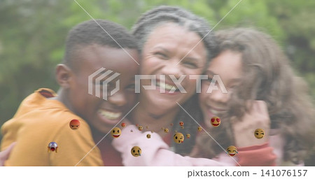 Smiling mother in red top and children leaning into hug at park, showing floating emoji reactions Smiling mother in red top and children leaning into hug at park, showing floating emoji reactions 141076157