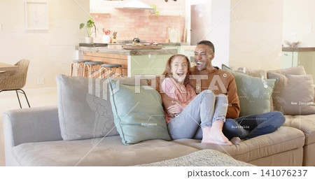 Sitting couple wearing brown sweater and pink hoodie, cuddling at home on sectional with dotted map Sitting couple wearing brown sweater and pink hoodie, cuddling at home on sectional with dotted map 141076237