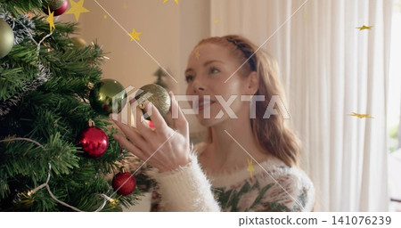 Standing woman in fuzzy holiday sweater placing gold bauble on Christmas tree at home, with lights Standing woman in fuzzy holiday sweater placing gold bauble on Christmas tree at home, with lights 141076239