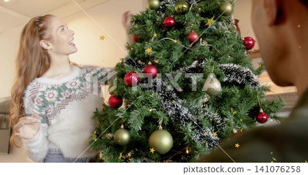 Smiling red-haired woman in white sweater placing ornament at home by decorated tree, man watching 141076258
