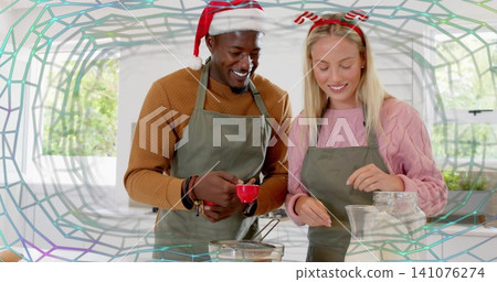 Measuring couple wearing aprons Santa hat antlers holding red cup, mixing in kitchen near flour jar Measuring couple wearing aprons Santa hat antlers holding red cup, mixing in kitchen near flour jar 141076274