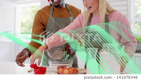 Mixing couple wearing aprons and sweaters at kitchen island, holding metal bowl, reaching eggs Mixing couple wearing aprons and sweaters at kitchen island, holding metal bowl, reaching eggs 141076346
