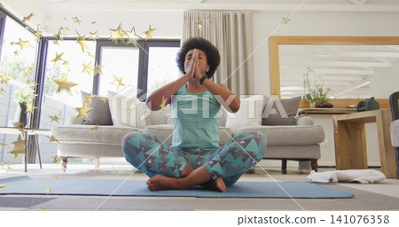 Meditating woman wearing teal outfit on blue yoga mat in living room with windows, gold stars Meditating woman wearing teal outfit on blue yoga mat in living room with windows, gold stars 141076358