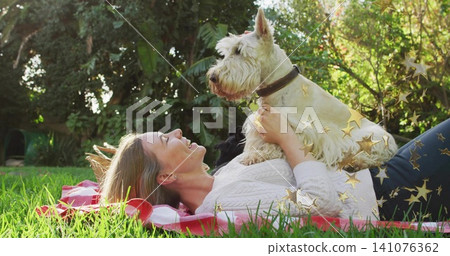 Lying woman holding white terrier on red-blanket in yard, wearing white-sweater darkpants goldstars Lying woman holding white terrier on red-blanket in yard, wearing white-sweater darkpants goldstars 141076362