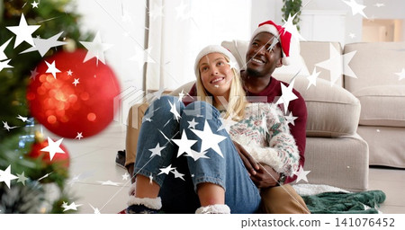 Embracing couple wearing Santa hat and knit beanie, cuddling on living room rug with red ornament Embracing couple wearing Santa hat and knit beanie, cuddling on living room rug with red ornament 141076452