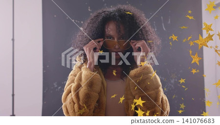 Pulling yellow aviator shades down, woman posing in studio with tan faux-fur coat, gold confetti 141076683