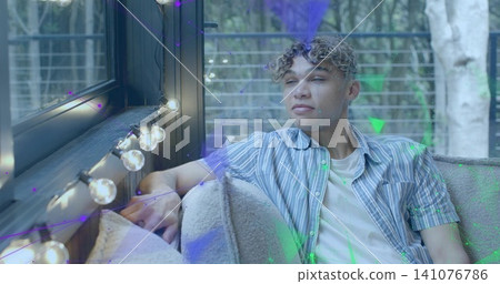 Reclining mid-20s male wearing striped shirt over white tee, resting arm on cushion by glass door Reclining mid-20s male wearing striped shirt over white tee, resting arm on cushion by glass door 141076786