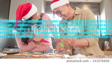 Decorating couple in Santa hats leaning over kitchen with binary band, piping red and green icing Decorating couple in Santa hats leaning over kitchen with binary band, piping red and green icing 141076809
