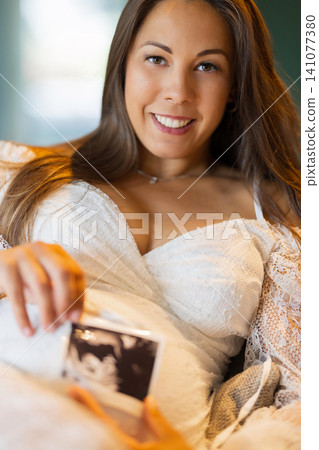 Pregnant woman smiling and holding an ultrasound photo at home Pregnant woman smiling and holding an ultrasound photo at home 141077380