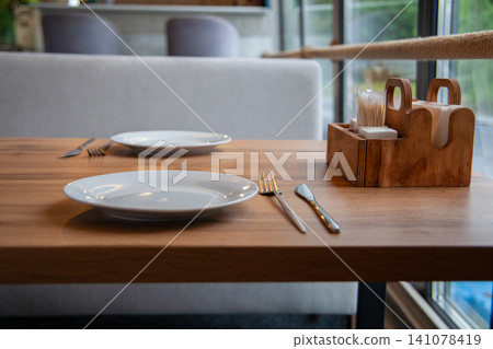 Close-up of wooden dining table set with empty plates indoor. Restaurant interior nobody Close-up of wooden dining table set with empty plates indoor. Restaurant interior nobody 141078419