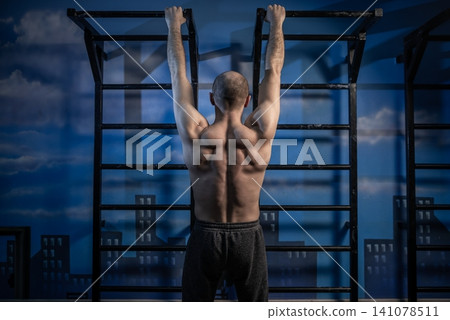 Rear view of a shirtless man hanging from overhead bars at a gym 141078511