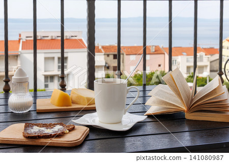 Breakfast table on a balcony in Makarska, Croatia, with coffee cup, open book, bread with butter, and Adriatic Sea view Breakfast table on a balcony in Makarska, Croatia, with coffee cup, open book, bread with butter, and Adriatic Sea view 141080987