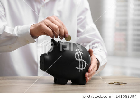 Man putting coin into piggy bank with dollar sign at wooden table, closeup Man putting coin into piggy bank with dollar sign at wooden table, closeup 141082394