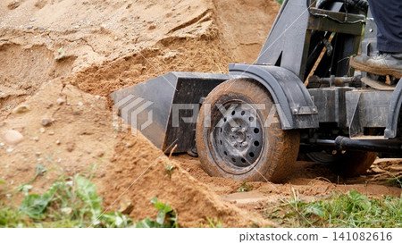 Close view of heavy construction machinery wheel moving through sandy excavation ground, with muddy metal parts, soil mounds, rough textures, and active earthmoving work outdoors 141082616