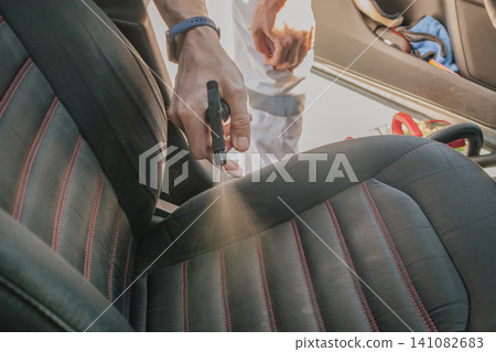 Horizontal close up shot of a person hand spraying chemical liquid cleaning detergent from a plastic bottle onto a black fabric car seat with red stitches Horizontal close up shot of a person hand spraying chemical liquid cleaning detergent from a plastic bottle onto a black fabric car seat with red stitches 141082683
