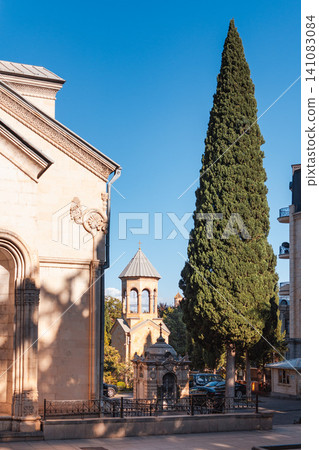 October 2024. Tbilisi, Georgia. Historic stone church with a conical dome and a tall green cypress tree under a clear blue sky. Vertical October 2024. Tbilisi, Georgia. Historic stone church with a conical dome and a tall green cypress tree under a clear blue sky. Vertical 141083084