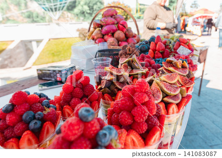 Market seller arranges assorted sliced fruit in transparent plastic cups at a street stand. Summer harvest concept highlights freshness, healthy eating, and local produce. Market seller arranges assorted sliced fruit in transparent plastic cups at a street stand. Summer harvest concept highlights freshness, healthy eating, and local produce. 141083087