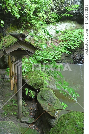 A votive tablet rack stands amidst the fresh greenery; a mystical early summer scene where moss-covered rocks, enshrined by the pond at Tsukimachi Falls, evoke a sense of history and faith. A votive tablet rack stands amidst the fresh greenery; a mystical early summer scene where moss-covered rocks, enshrined by the pond at Tsukimachi Falls, evoke a sense of history and faith. 141083825