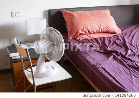 A white fan is positioned on a chair next to a bedside table and a rumpled bed with purple sheets and an orange pillow. 141084266