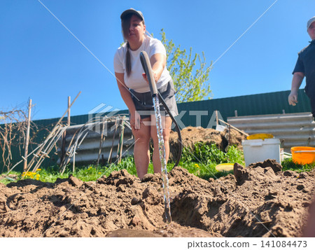 Woman in a baseball cap and t-shirt watering freshly tilled brown soil in a home garden on a sunny spring day, preparing the ground for planting new crops and nurturing growth. Woman in a baseball cap and t-shirt watering freshly tilled brown soil in a home garden on a sunny spring day, preparing the ground for planting new crops and nurturing growth. 141084473