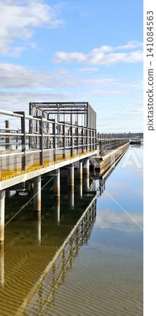 A modern metal pier extending into the mirror-like surface of the lake under a blue sky 141084563