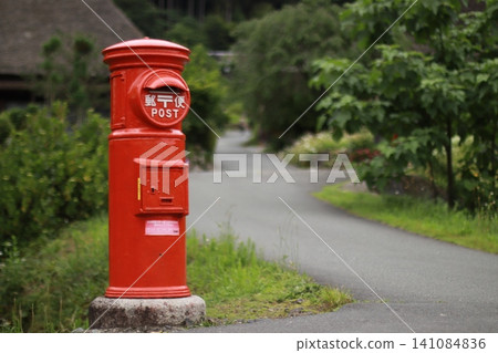 A red round mailbox stands beside a thatched-roof house. A red round mailbox stands beside a thatched-roof house. 141084836