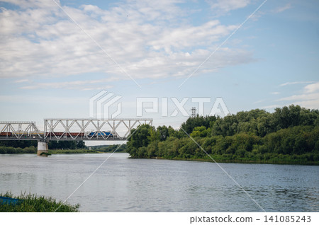 train crossing bridge over serene river landscape with trees and sky from riverbank perspective, modern transportation infrastructure with metal, concrete, and wood materials, vivid blue. 141085243