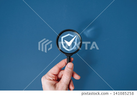 Hand holding magnifying glass over protective shield icon on blue background, data security and insurance concept Hand holding magnifying glass over protective shield icon on blue background, data security and insurance concept 141085258