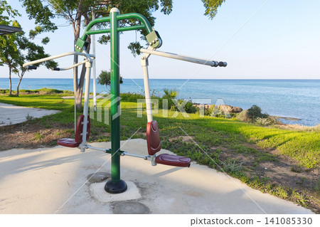 Outdoor fitness equipment overlooking the sea in a coastal park. Public exercise machine by the waterfront surrounded by green trees, offering a scenic place for healthy recreation. 141085330