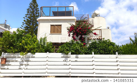Modern white Mediterranean house with lush garden and blooming bougainvillea under a bright blue sky. Cozy residential architecture in Cyprus with vibrant summer atmosphere. Modern white Mediterranean house with lush garden and blooming bougainvillea under a bright blue sky. Cozy residential architecture in Cyprus with vibrant summer atmosphere. 141085331