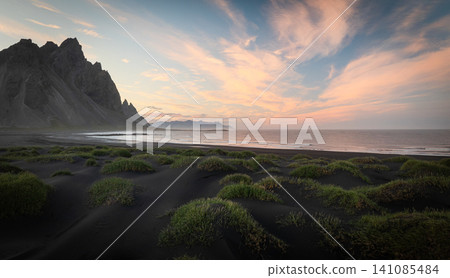 Colorful sunset over the iconic Vestrahorn mountain with black volcanic sand dunes and the Atlantic coastline. Peaceful Icelandic landscape with dramatic sky and untouched nature. 141085484