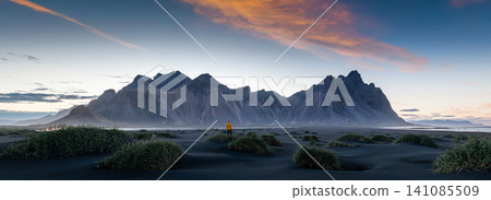Wide panoramic view of Vestrahorn at sunset with a lone traveler standing on Iceland's black sand beach. Dramatic mountains, colorful clouds and volcanic landscape create an epic scene. Wide panoramic view of Vestrahorn at sunset with a lone traveler standing on Iceland's black sand beach. Dramatic mountains, colorful clouds and volcanic landscape create an epic scene. 141085509