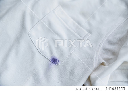 Close-up of a purple ink stain from a pen on the pocket of a white polo shirt. Concept of dirty clothes and laundry stain removal. Close-up of a purple ink stain from a pen on the pocket of a white polo shirt. Concept of dirty clothes and laundry stain removal. 141085555