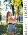 Teenager girl enjoying nature in a park during a sunny autumn day, touching colorful leaves on a branch 141086404