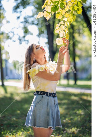 Teenager girl enjoying nature in a park during a sunny autumn day, touching colorful leaves on a branch Teenager girl enjoying nature in a park during a sunny autumn day, touching colorful leaves on a branch 141086404