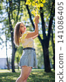 Young woman smiling, wearing trendy outfit, reaching up to colorful autumn foliage during sunny park afternoon 141086405
