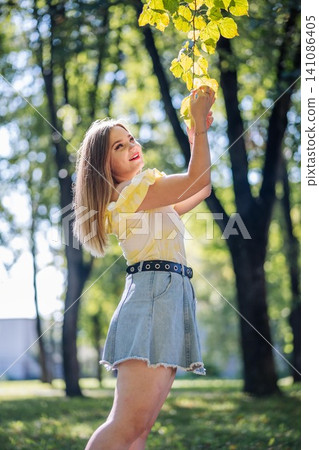 Young woman smiling, wearing trendy outfit, reaching up to colorful autumn foliage during sunny park afternoon Young woman smiling, wearing trendy outfit, reaching up to colorful autumn foliage during sunny park afternoon 141086405