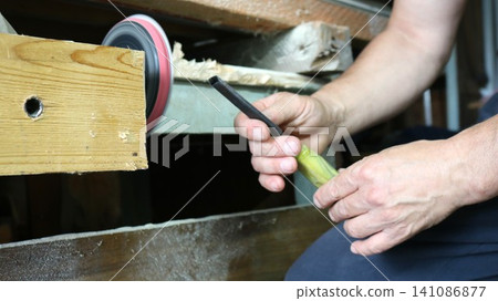A craftsman sharpens a chisel in a woodworking shop. The man's hands carefully guide the tool's metal blade to the rotating grinding wheel of a sharpening device near a wooden workbench. 141086877