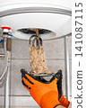 Plumber Holding Removed Water Heater Heating Element Covered With Heavy Mineral Deposits, Showing Hard Water Damage And Need For Maintenance Service 141087115
