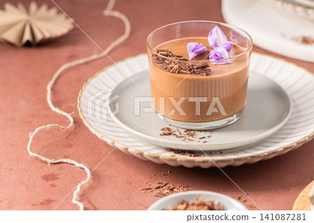 A single glass of chocolate mousse, beautifully garnished with chocolate shavings and purple flowers on a plate, styled with rustic elements. A single glass of chocolate mousse, beautifully garnished with chocolate shavings and purple flowers on a plate, styled with rustic elements. 141087281