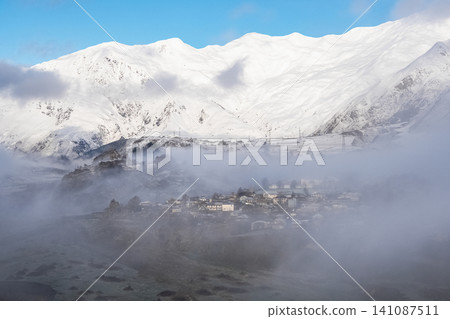 Scenic view of a mountain village surrounded by heavy morning fog and snow-covered peaks, Georgia Scenic view of a mountain village surrounded by heavy morning fog and snow-covered peaks, Georgia 141087511