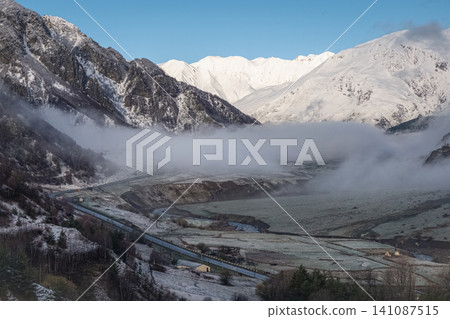 Scenic winter landscape of Caucasus mountains in Georgia with mountain road, river, and thick low clouds. Scenic winter landscape of Caucasus mountains in Georgia with mountain road, river, and thick low clouds. 141087515