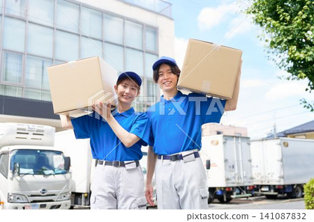 A male and female delivery staff member smiles as they carry cardboard boxes on their shoulders. 141087832