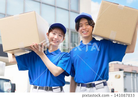 Young delivery workers in uniform carrying cardboard boxes outdoors. Young delivery workers in uniform carrying cardboard boxes outdoors. 141087833