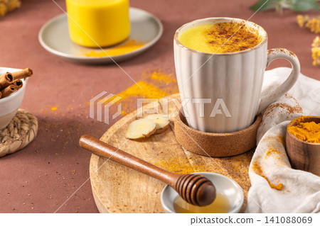 A warm and creamy golden milk latte in a white ribbed mug surrounded by fresh ginger root turmeric powder cinnamon and honey for a cozy holistic wellness drink. Vibrant Healthy Golden Milk Turmeric A warm and creamy golden milk latte in a white ribbed mug surrounded by fresh ginger root turmeric powder cinnamon and honey for a cozy holistic wellness drink. Vibrant Healthy Golden Milk Turmeric 141088069