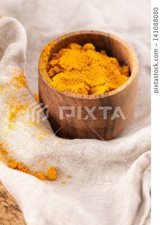 Ground turmeric spice fills a small wooden bowl resting on a textured linen cloth with scattered yellow powder highlights for a natural kitchen aesthetic Ground turmeric spice fills a small wooden bowl resting on a textured linen cloth with scattered yellow powder highlights for a natural kitchen aesthetic 141088080