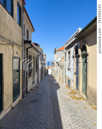 A sun-drenched, narrow cobblestone lane in Porto leads toward the sparkling blue Atlantic Ocean, flanked by historic houses and charming vintage doors A sun-drenched, narrow cobblestone lane in Porto leads toward the sparkling blue Atlantic Ocean, flanked by historic houses and charming vintage doors 141088143