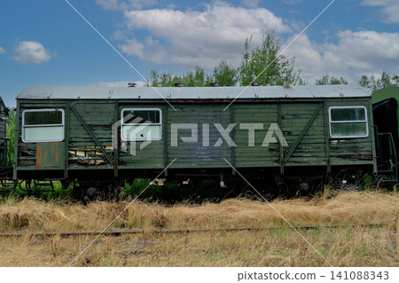 A weathered green wooden train carriage with peeling paint standing on dry grass under cloudy sky A weathered green wooden train carriage with peeling paint standing on dry grass under cloudy sky 141088343